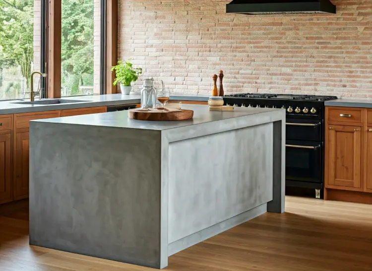 A cozy kitchen with a large concrete island countertop, exposed brick walls, wooden cabinets in a rustic style, and an airy concrete benchtop