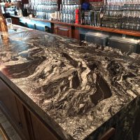 A marble-topped bar counter with numerous wine bottles, featuring Black Forest Leather Granite and Venatino Quantum Quartz.